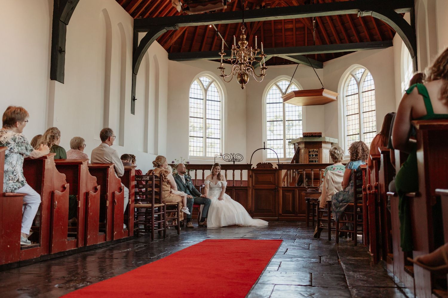 Bride in white gown walks down red carpet aisle in historic white church with wooden pews and chandelier.
