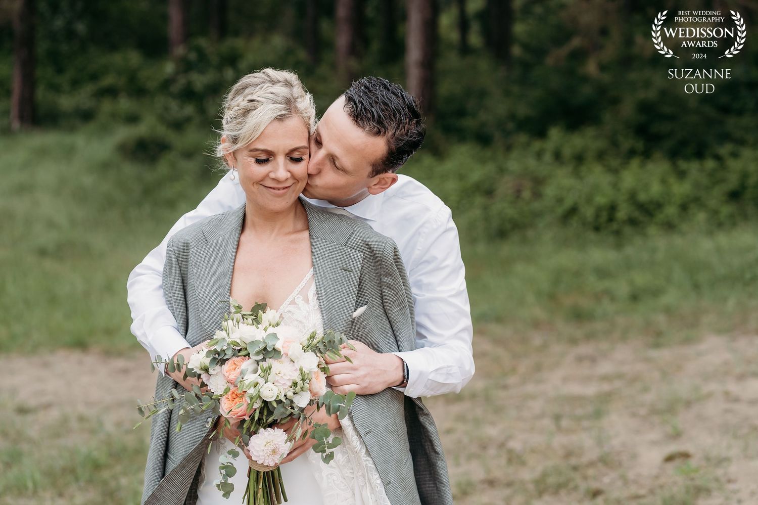 A couple embraces in a romantic moment outdoors, with the bride holding a peach and white floral bouquet.