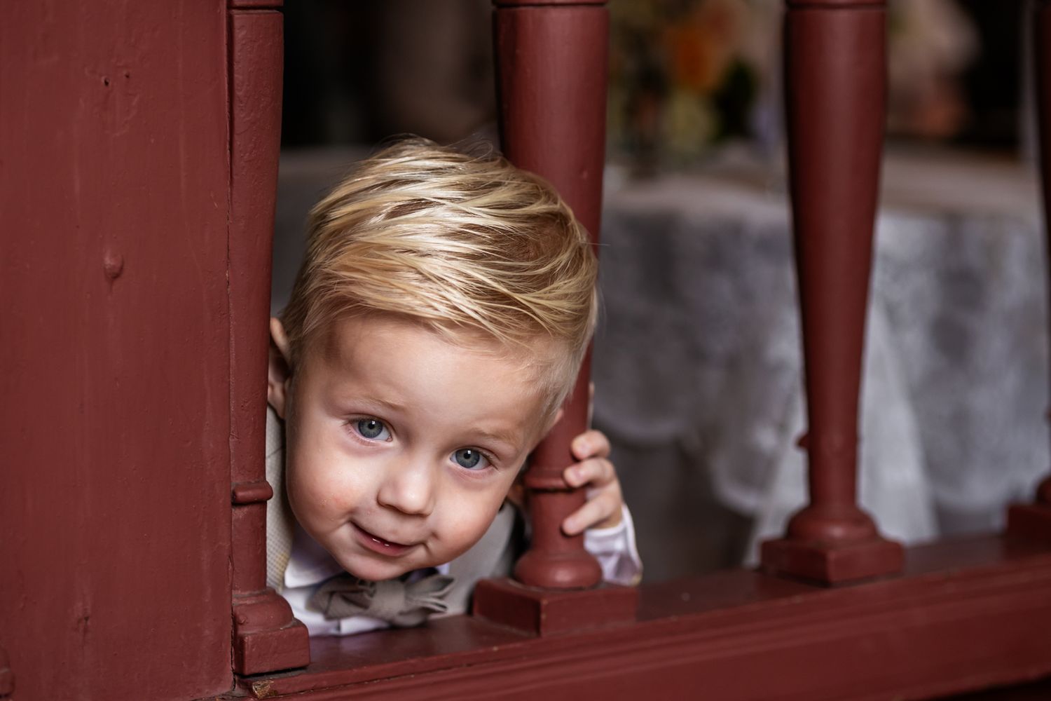 Young child peeks through red wooden railings during wedding ceremony.