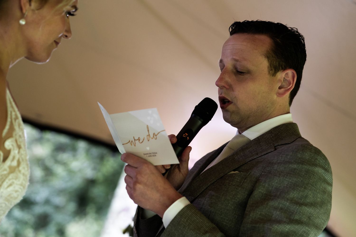 Wedding ceremony moment showing someone reading from papers while holding a microphone under a tent.