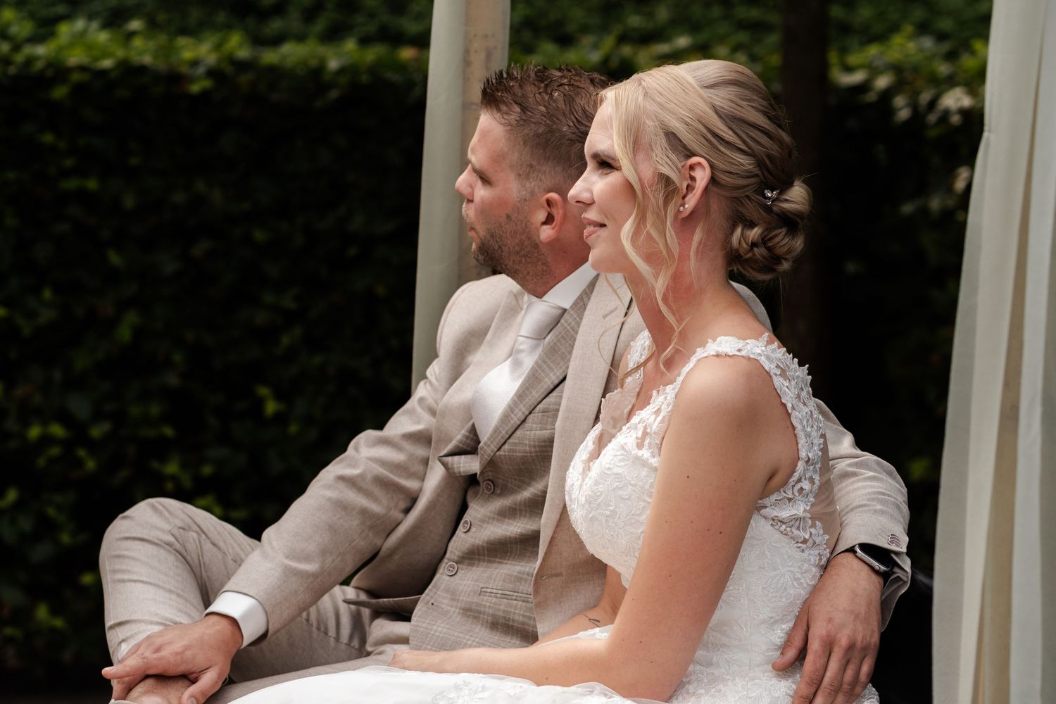 A happy couple sits together under a gazebo wearing elegant wedding attire on a sunny day.