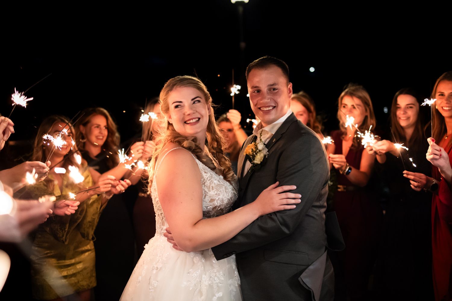 Newlyweds dancing while wedding guests hold sparklers during nighttime celebration.