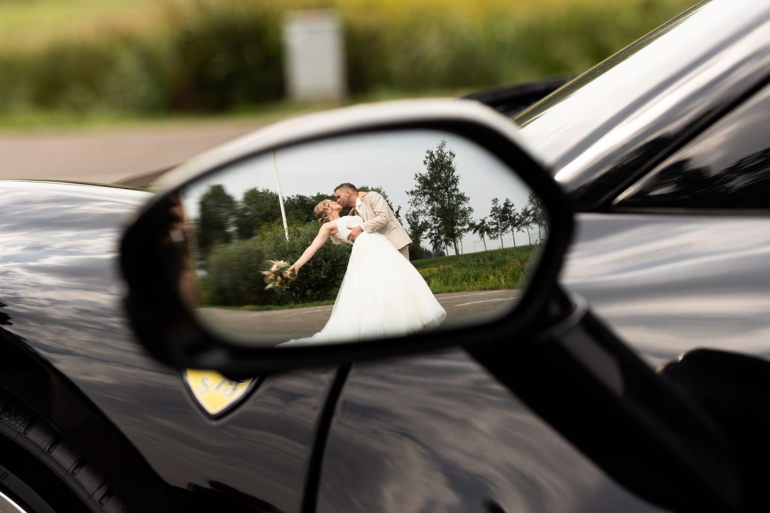 Creative wedding photo showing a couple reflected in a black car's side mirror against a scenic landscape.