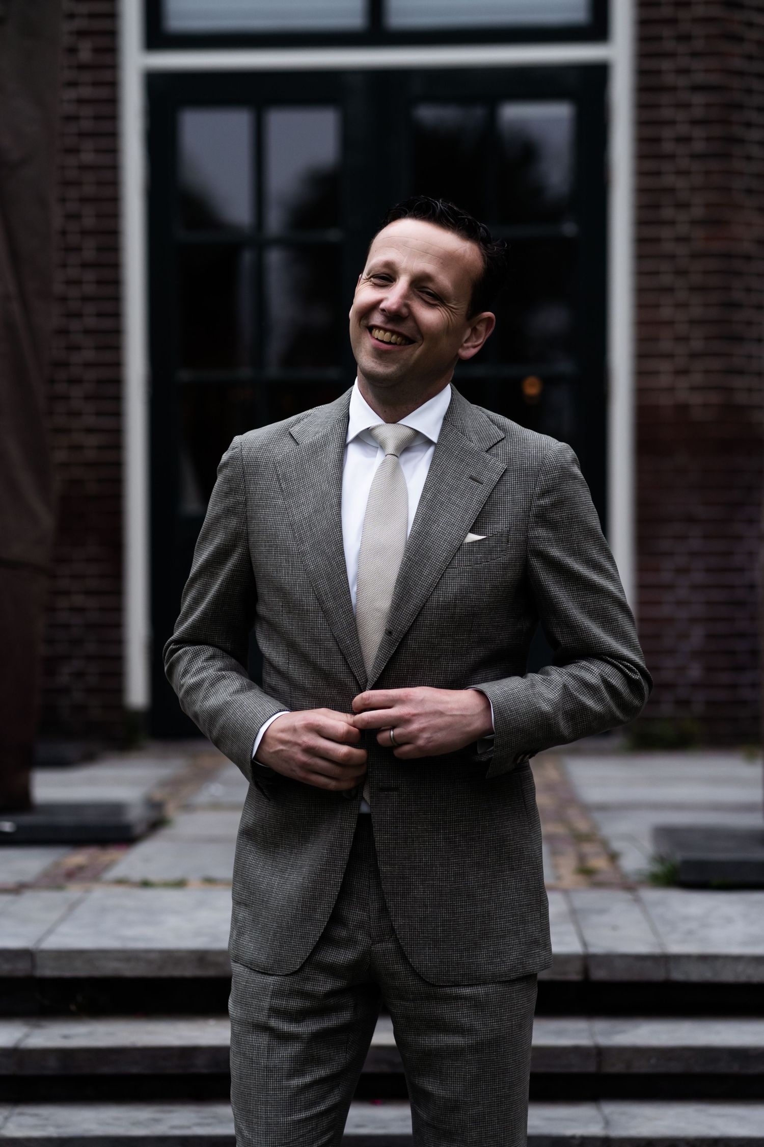 Person in grey suit smiles while standing outside brick building.