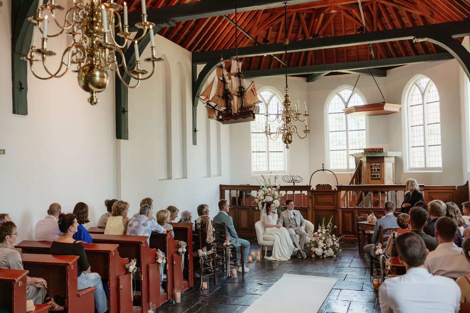 Wedding ceremony in a historic white Dutch church with red pews, chandeliers, and arched windows, guests seated as couple stands at altar.