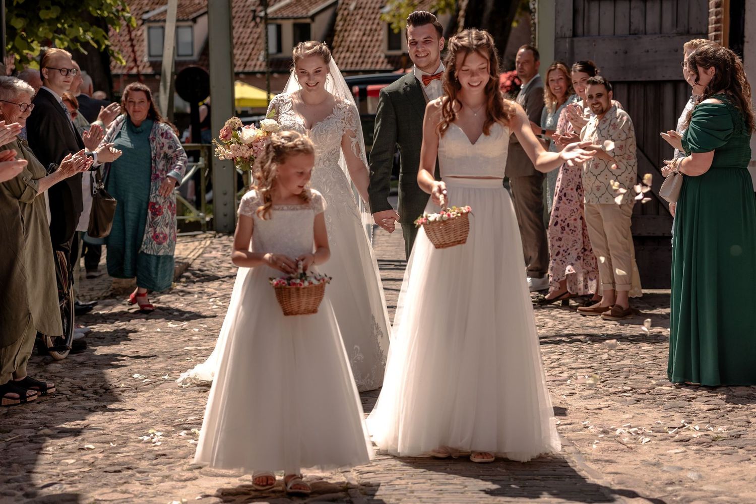 Flower girls in white dresses hold woven baskets during an outdoor wedding ceremony.