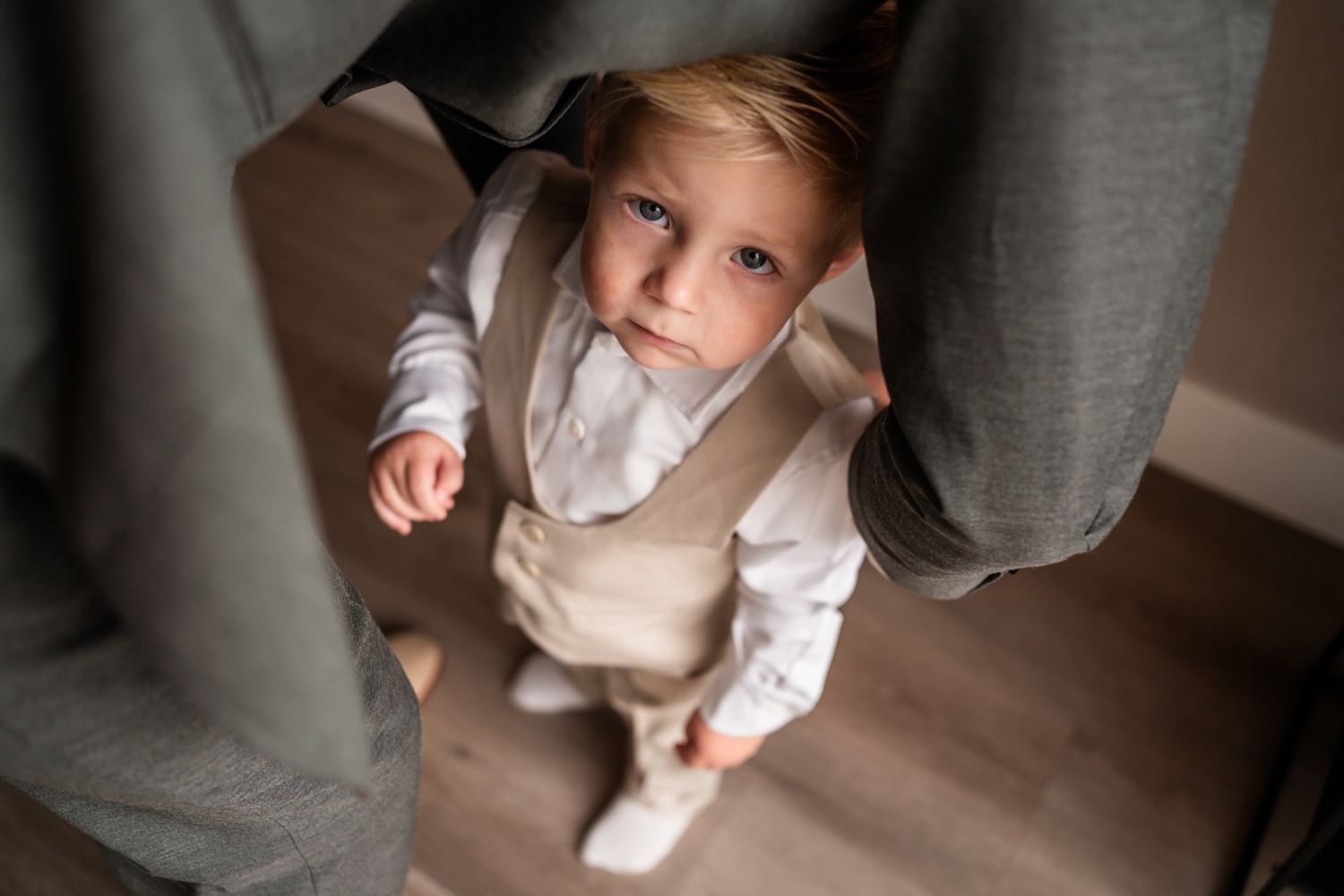Small child in formal beige outfit and white shirt standing between curtains indoors.