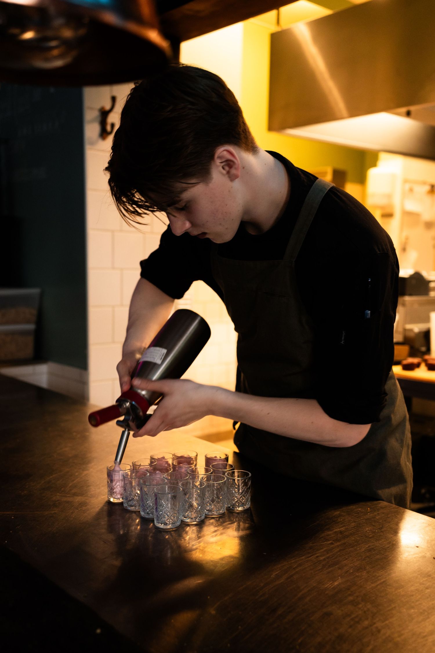 Bartender pouring drinks in dimly lit bar with multiple shot glasses lined up on dark wooden counter.