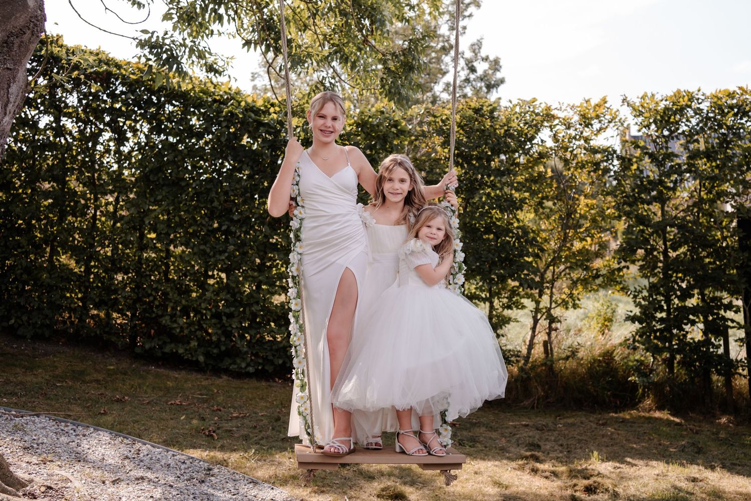 Two people in elegant white dresses share a playful moment in a garden setting with lush greenery.