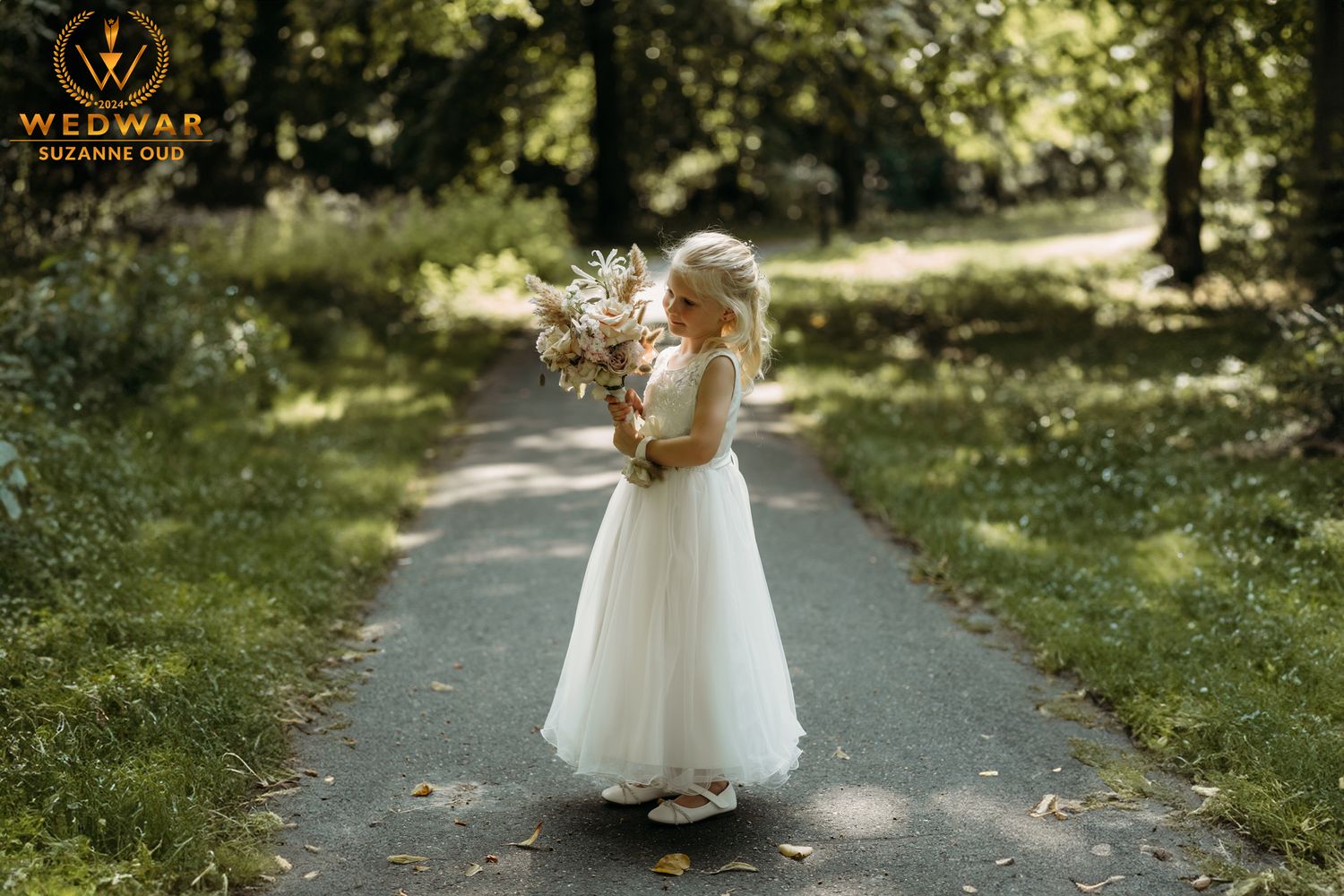 Young attendant in white dress holding bouquet on tree-lined path.