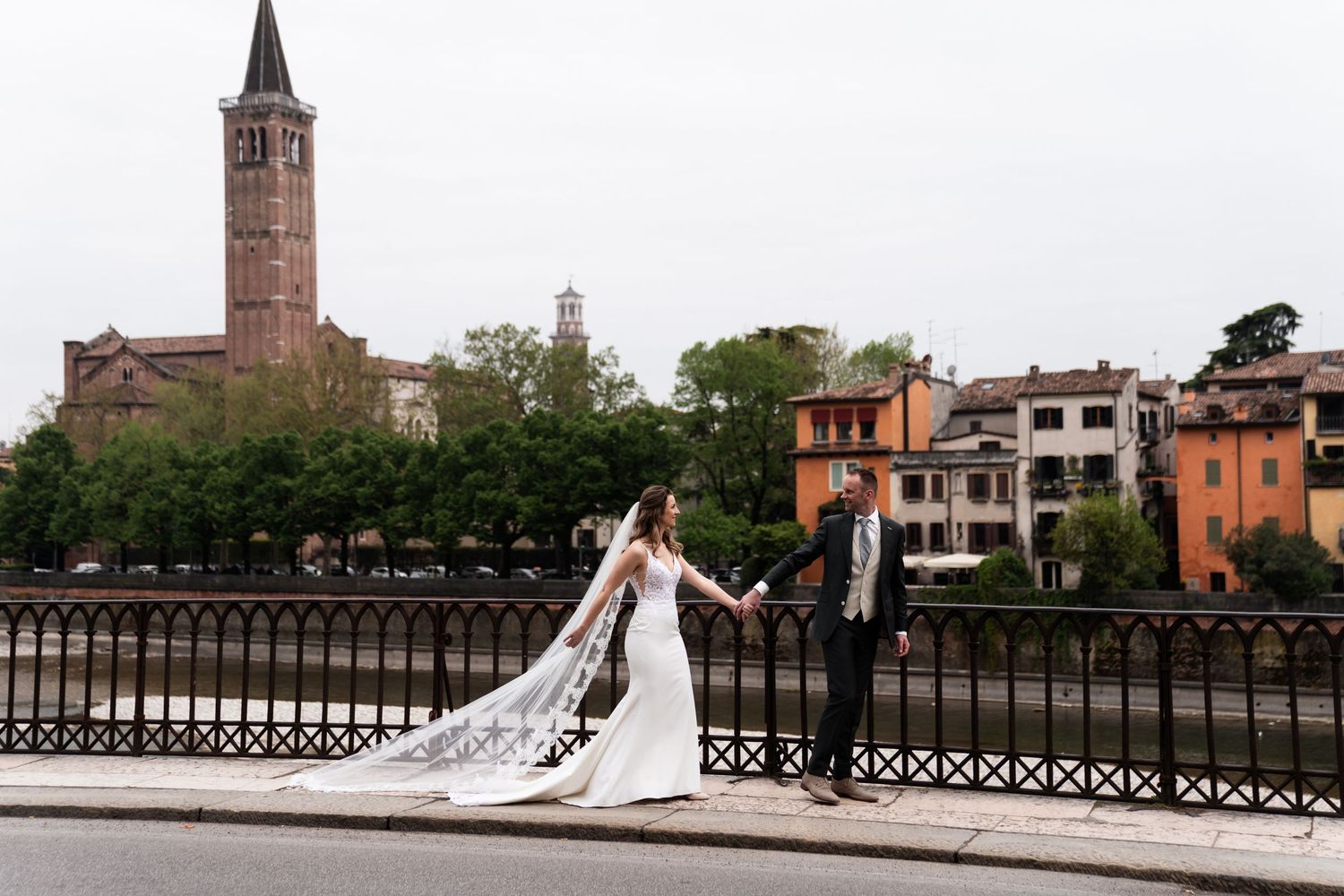 A couple in wedding attire poses on a bridge in Verona, Italy with historic architecture and church spire in background.
