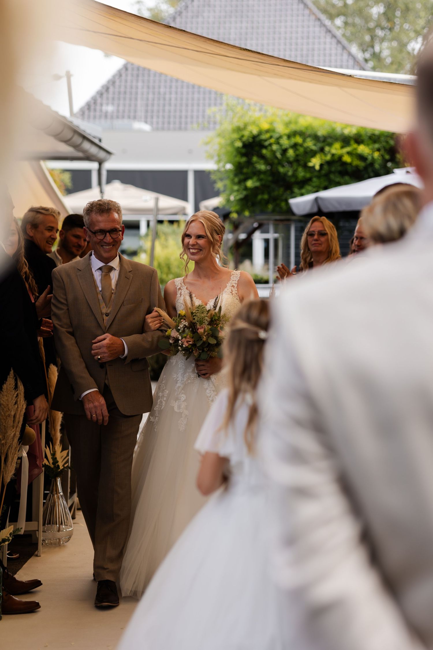 Wedding ceremony processional with bride in white gown holding floral bouquet walking down outdoor covered aisle.