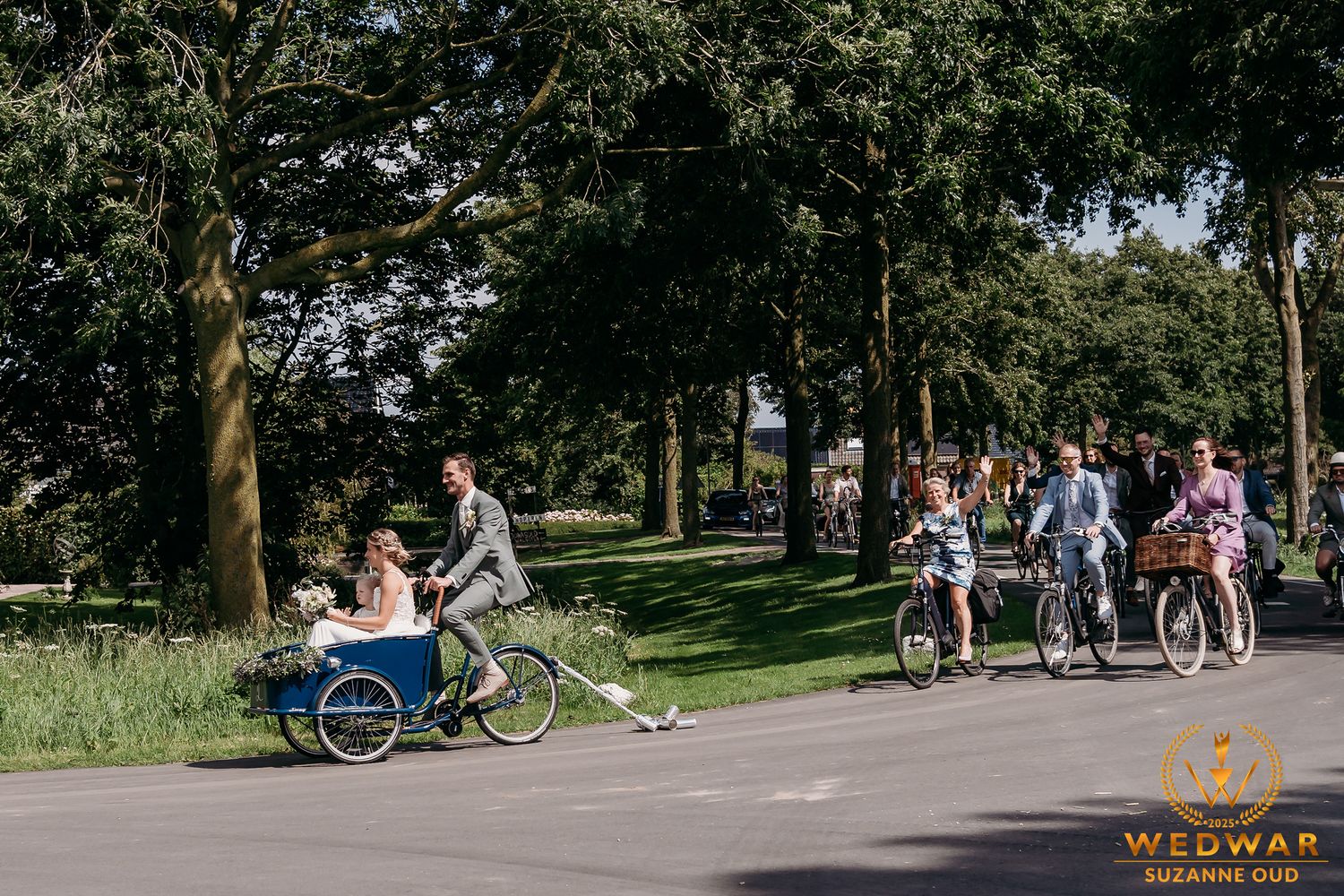 People enjoying a sunny day in a park with bikes and strollers on a paved path surrounded by green trees.
