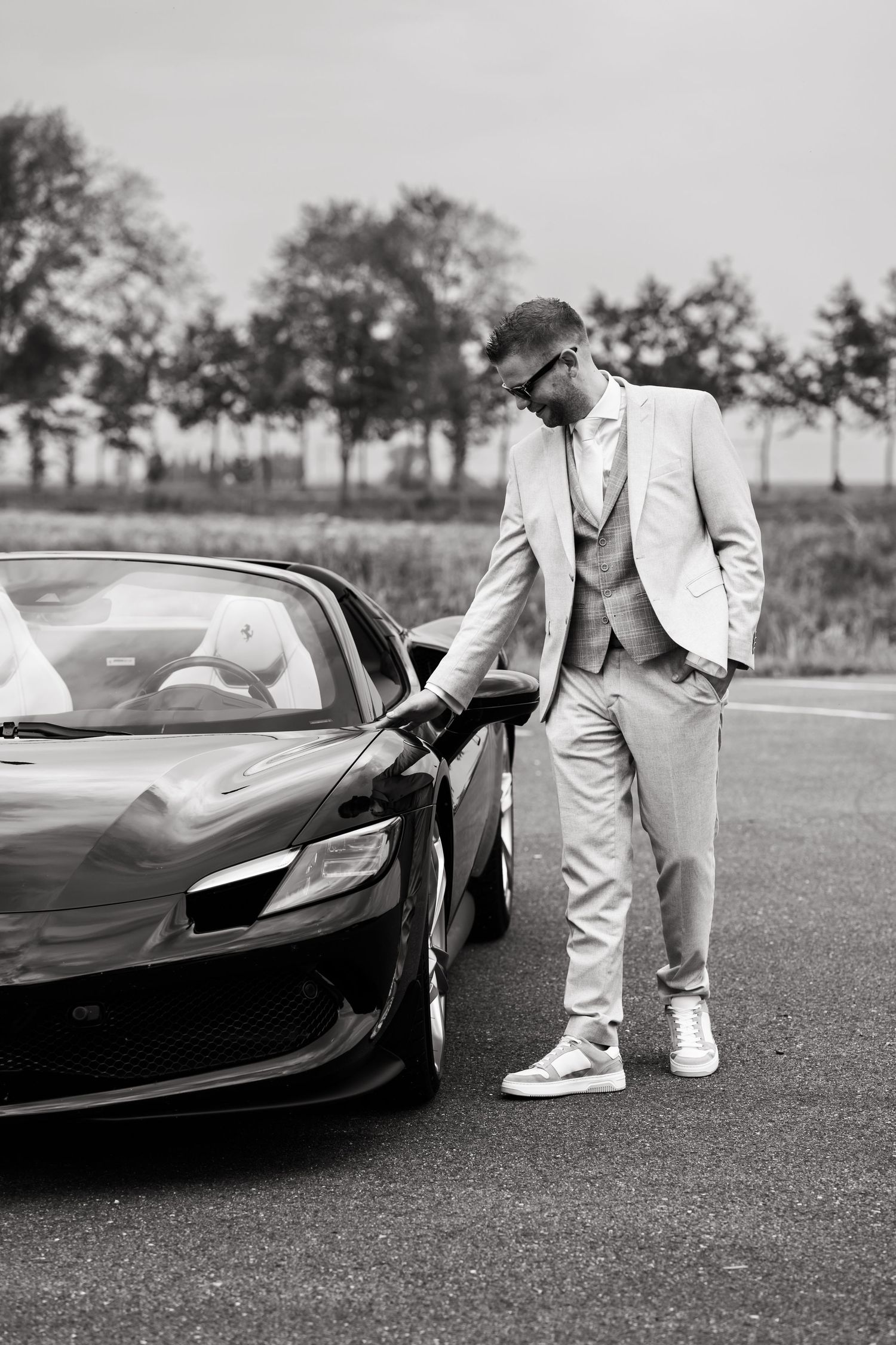 A stylishly dressed person in a light suit poses next to a black luxury sports car.