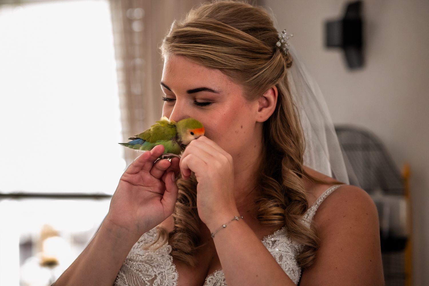 Person in a white wedding dress smells a green budgie perched on their finger.