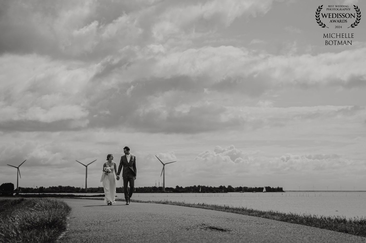 Black and white silhouette of couple walking along path with wind turbines in background.