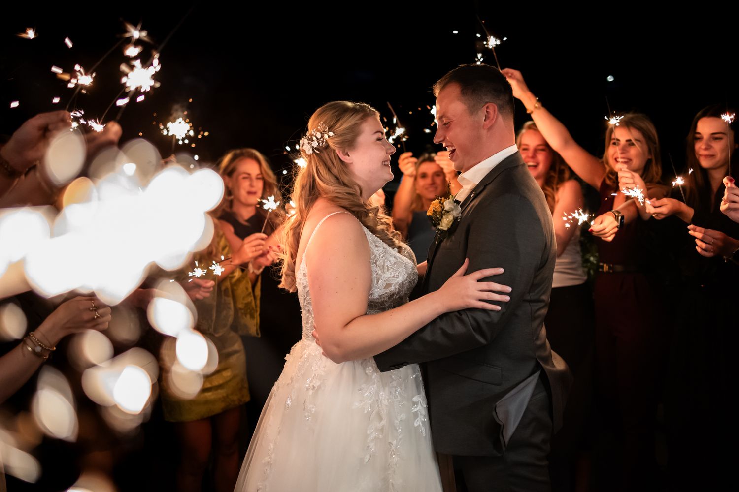 A bride and groom share their first dance surrounded by sparklers and wedding guests in a romantic nighttime setting.
