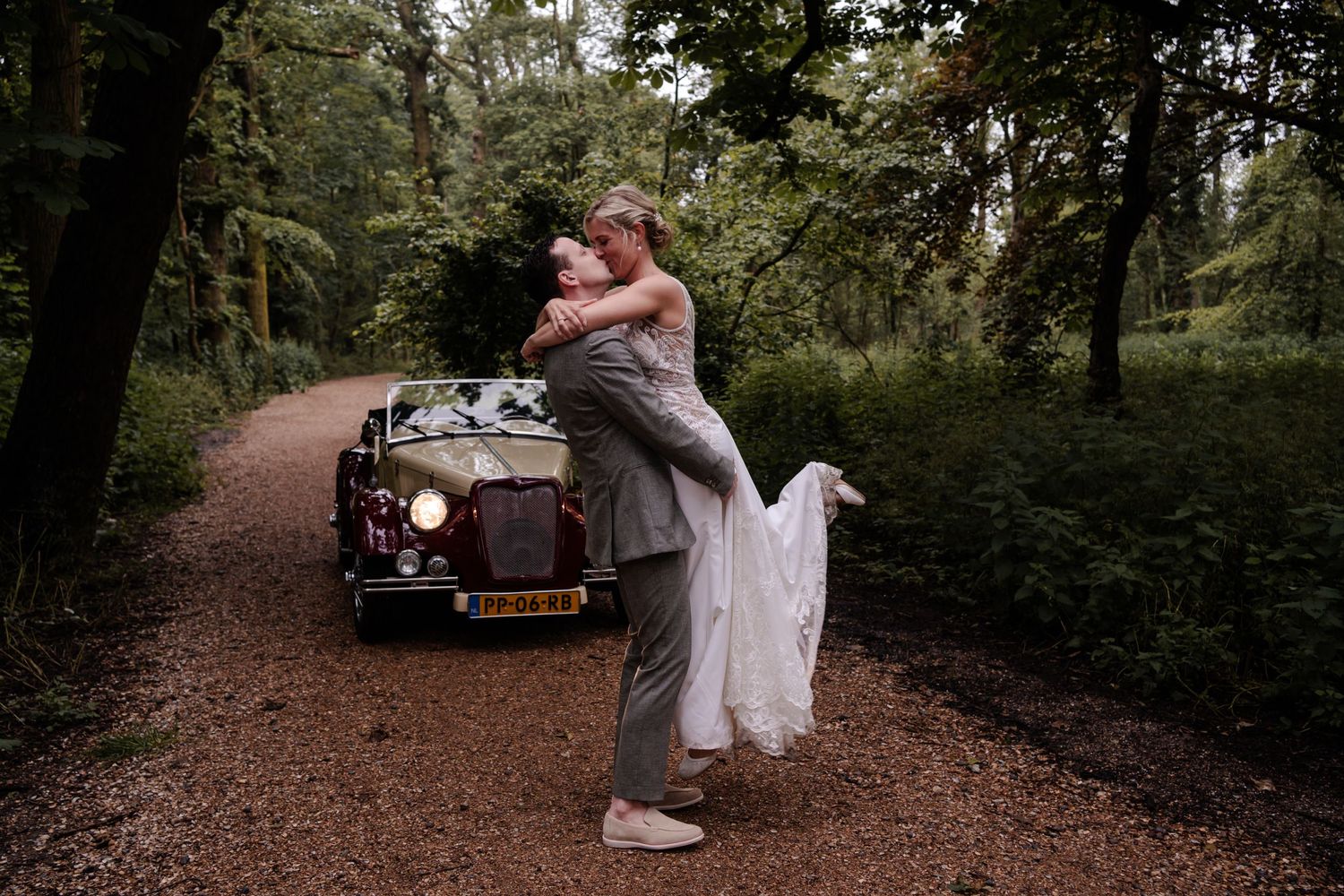 A couple embraces in front of a classic vintage car on a forest path.