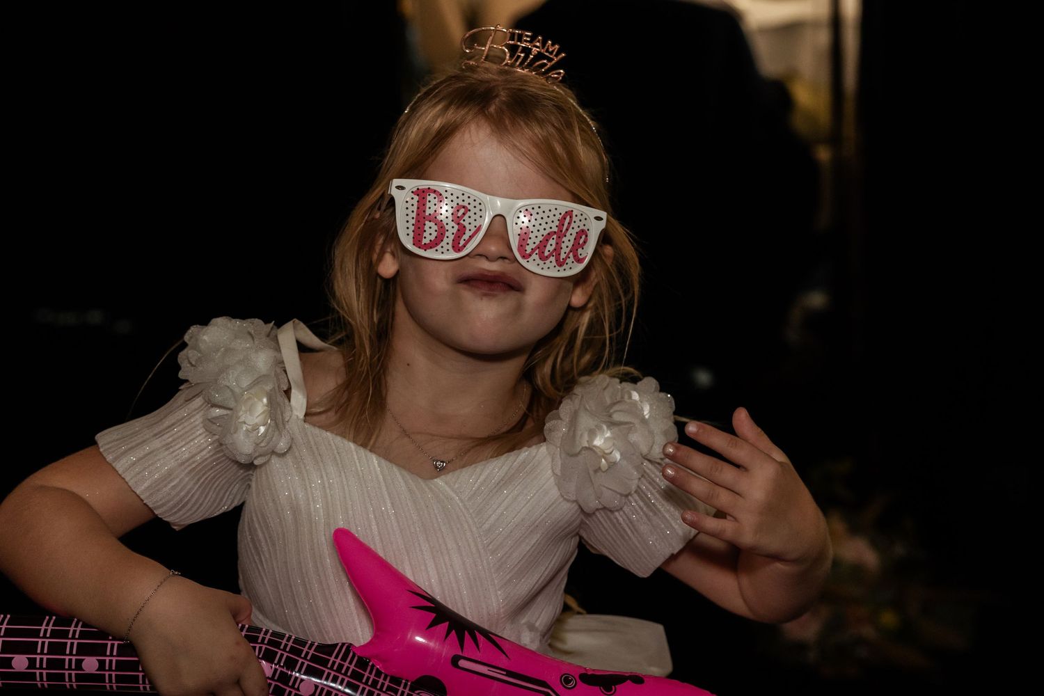 Child wearing a white dress and blindfold mask holds a pink toy at a party.