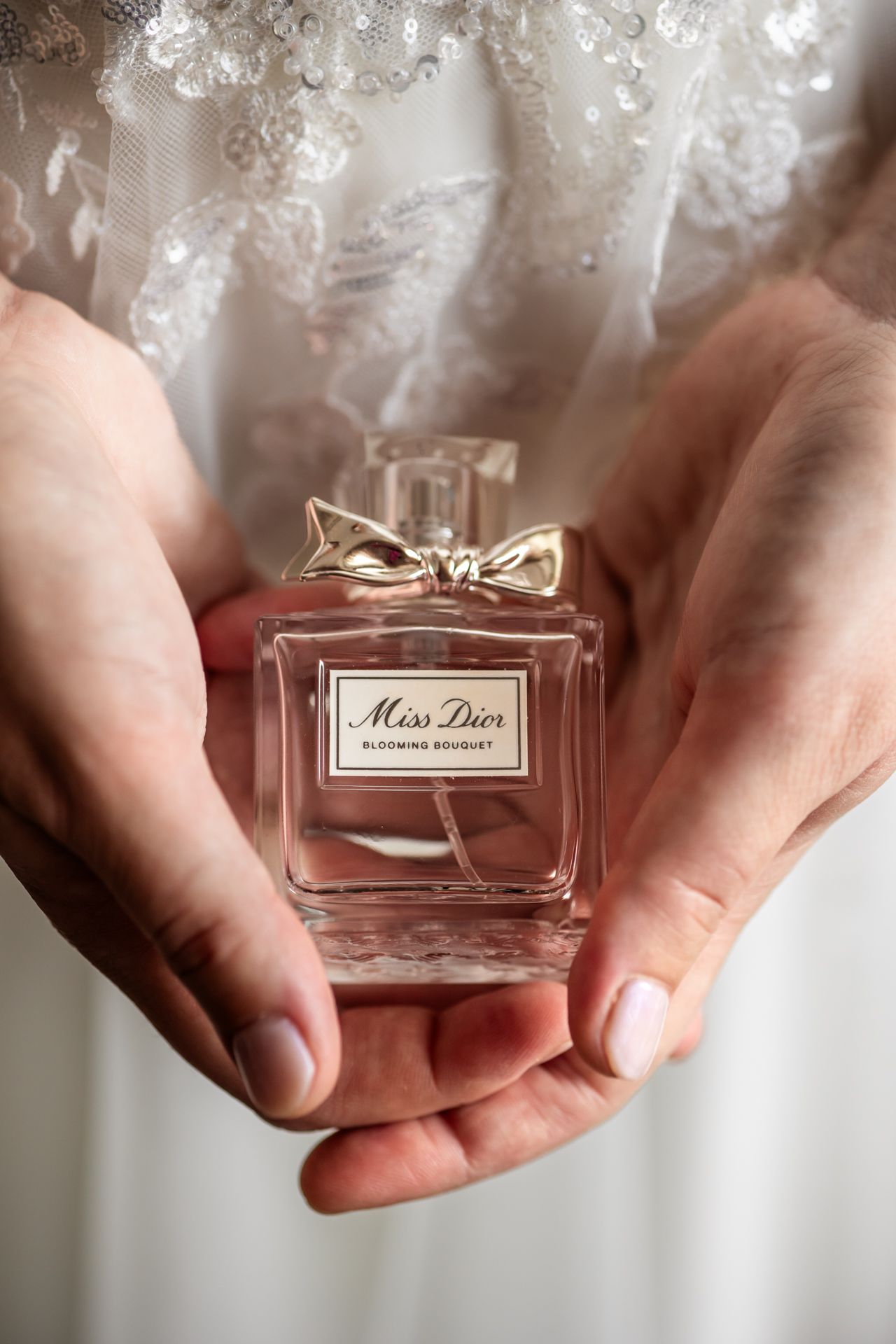 Hands gently hold a delicate bottle of Miss Dior perfume against a soft white lace background.