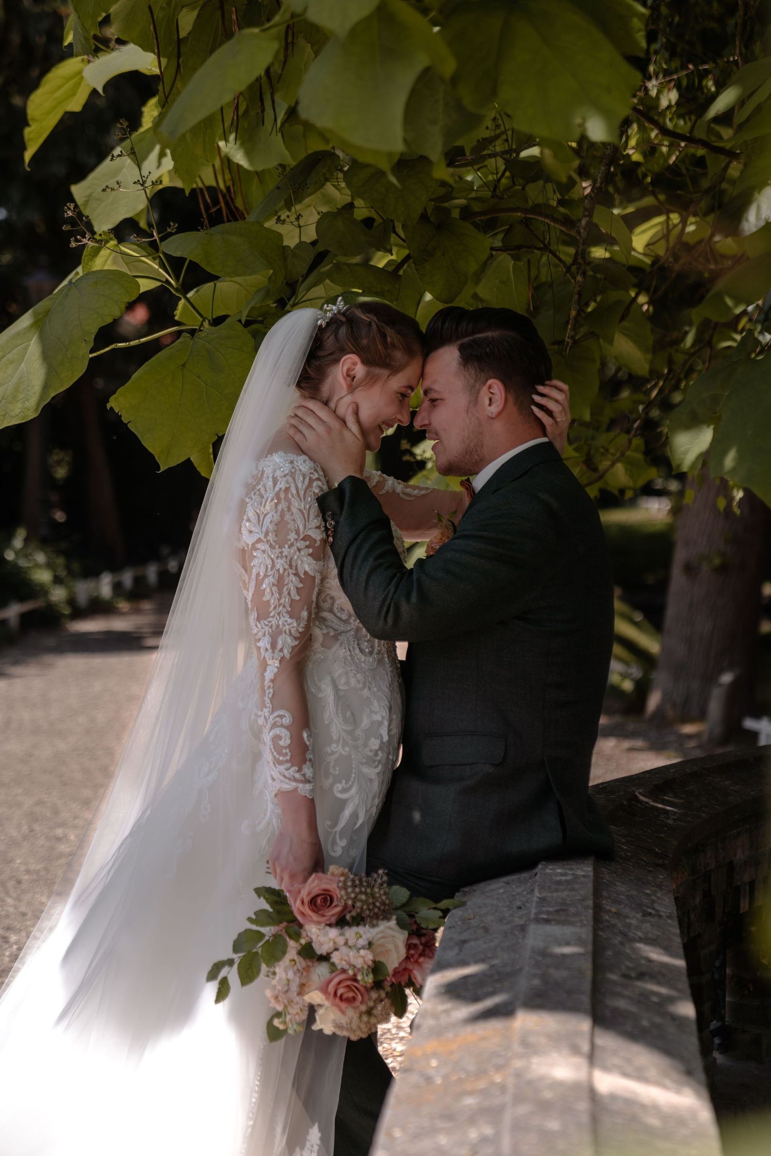 A romantic wedding couple shares intimate moments under green foliage in a garden setting.