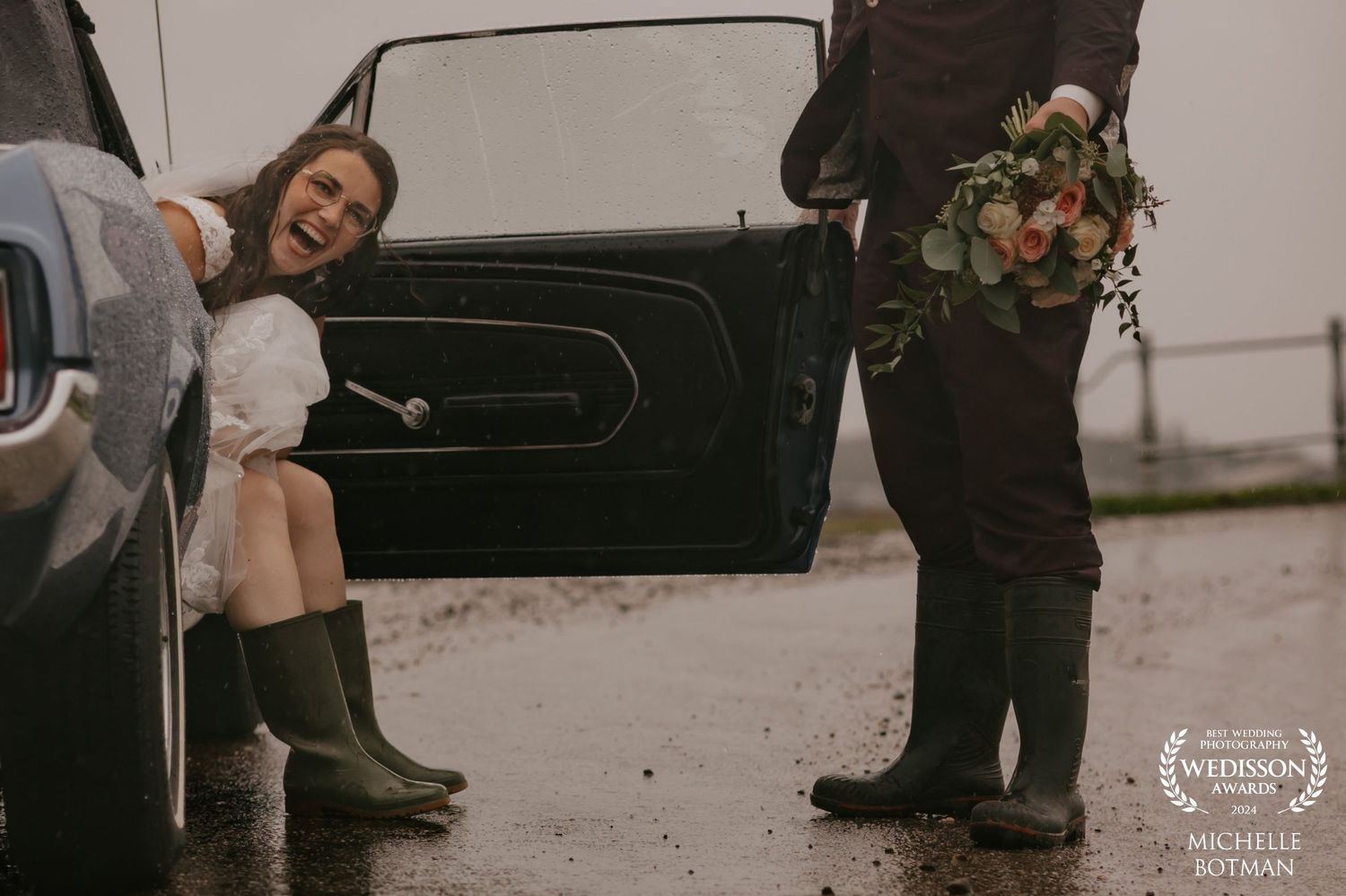 Candid moment with wellington boots beside vintage car on rainy wedding day.