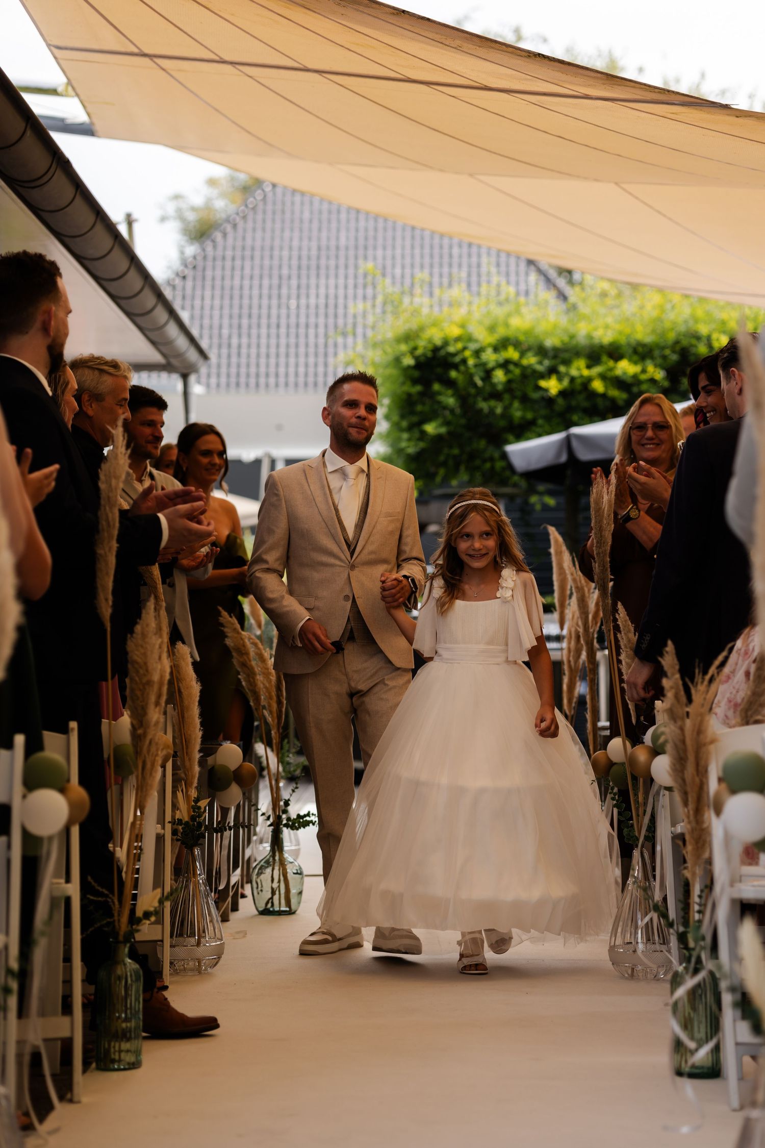 Wedding ceremony processional on an outdoor covered walkway adorned with lights and lined with guests on either side.