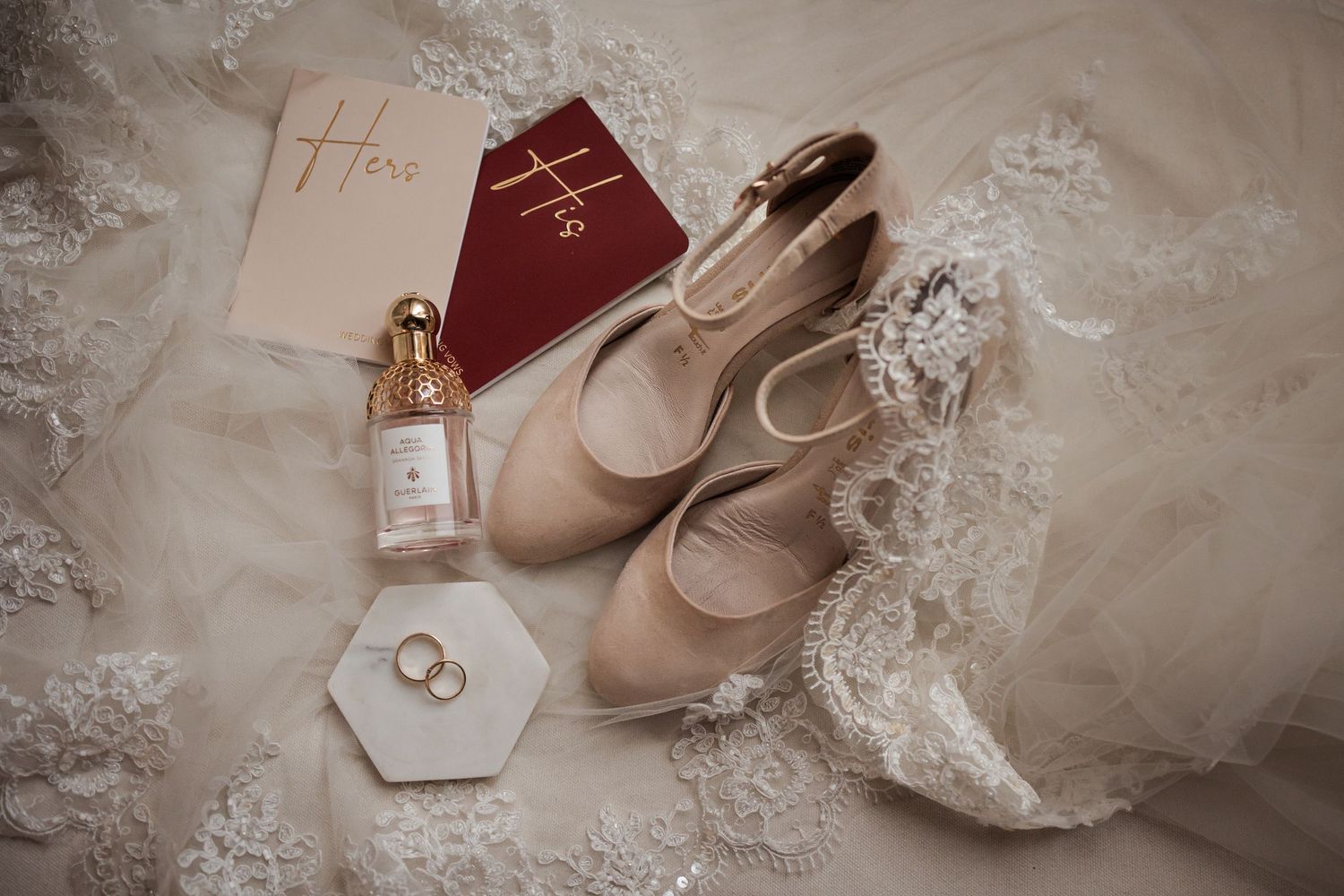 Wedding accessories including lace veil, perfume bottle, ballet flats, and delicate jewelry arranged on light fabric.