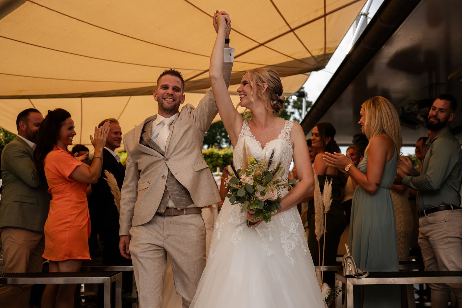 A couple celebrates joyfully under a beige tent at an outdoor wedding reception with guests cheering in the background.