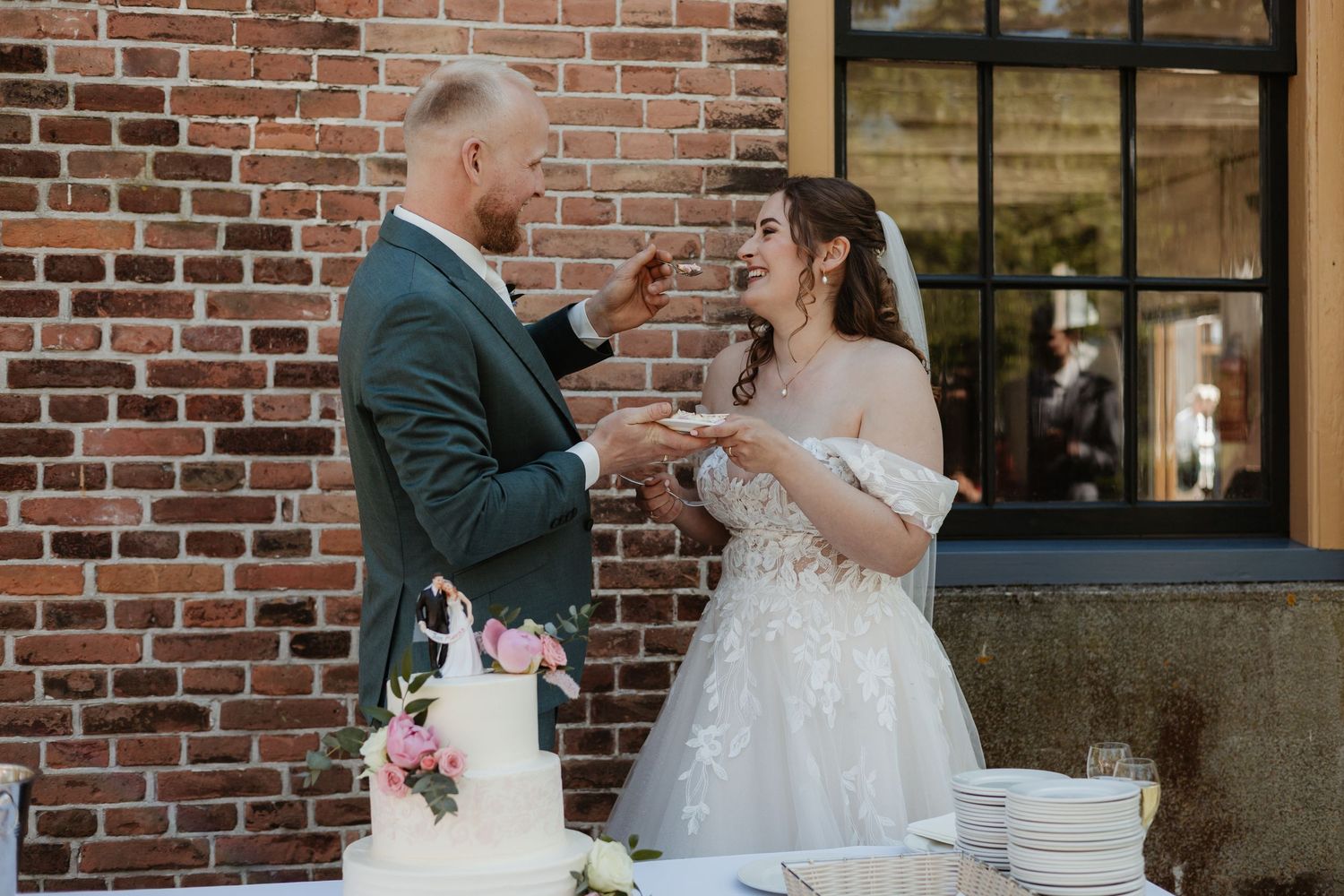 Bride and groom laughing while cutting their white wedding cake outdoors against a brick wall backdrop.