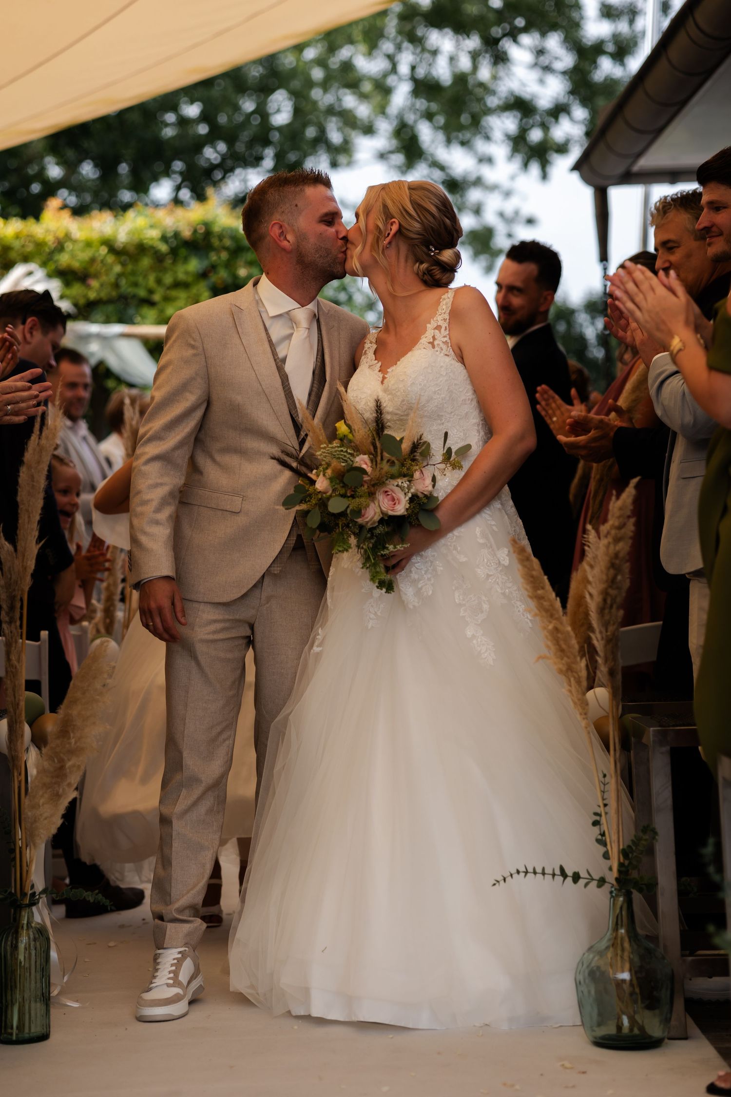A couple shares a tender kiss at their outdoor wedding ceremony while wearing elegant attire and holding a bouquet.