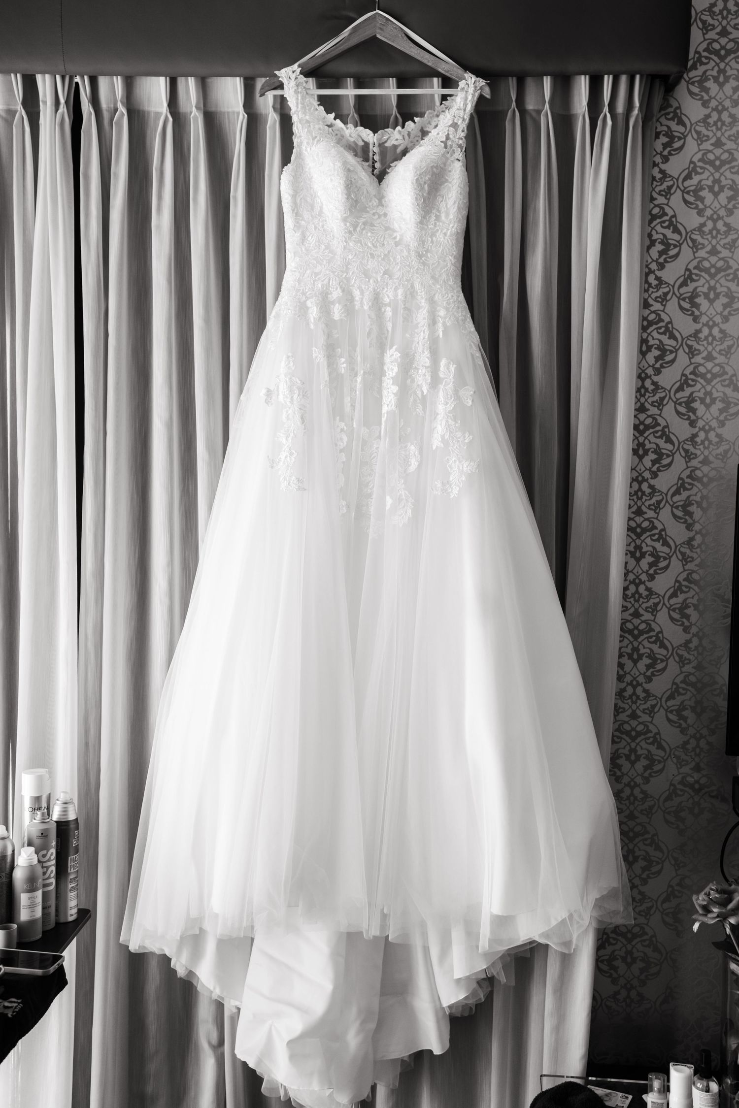 A classic white wedding dress with lace bodice and tulle skirt hangs elegantly against curtains in black and white.