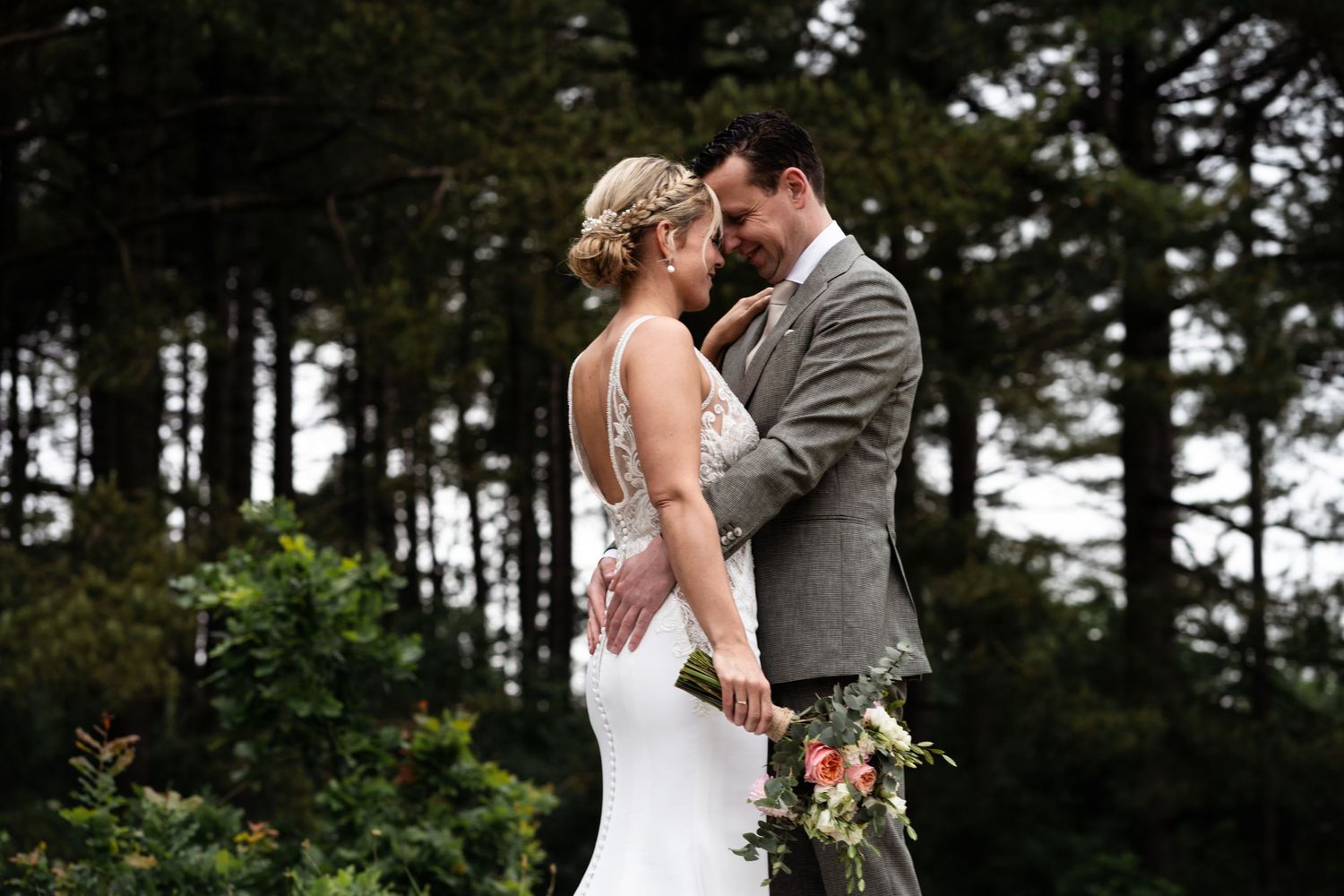 Two people share a romantic moment in their wedding attire against a backdrop of evergreen trees.