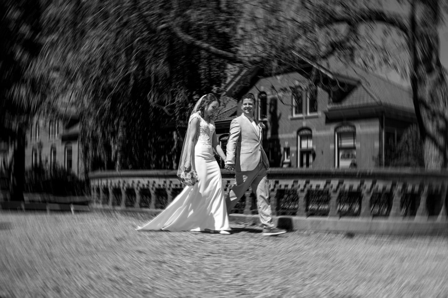 Black and white photo of a bride and groom walking hand in hand on a cobblestone path near a historic mansion.
