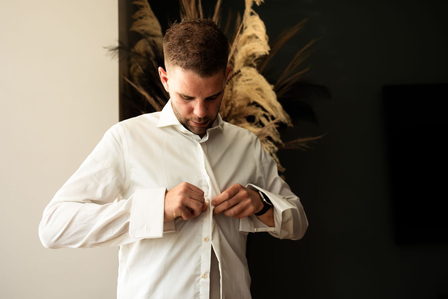 Someone in a white button-up shirt fastening buttons against a dark background with decorative dried pampas grass.