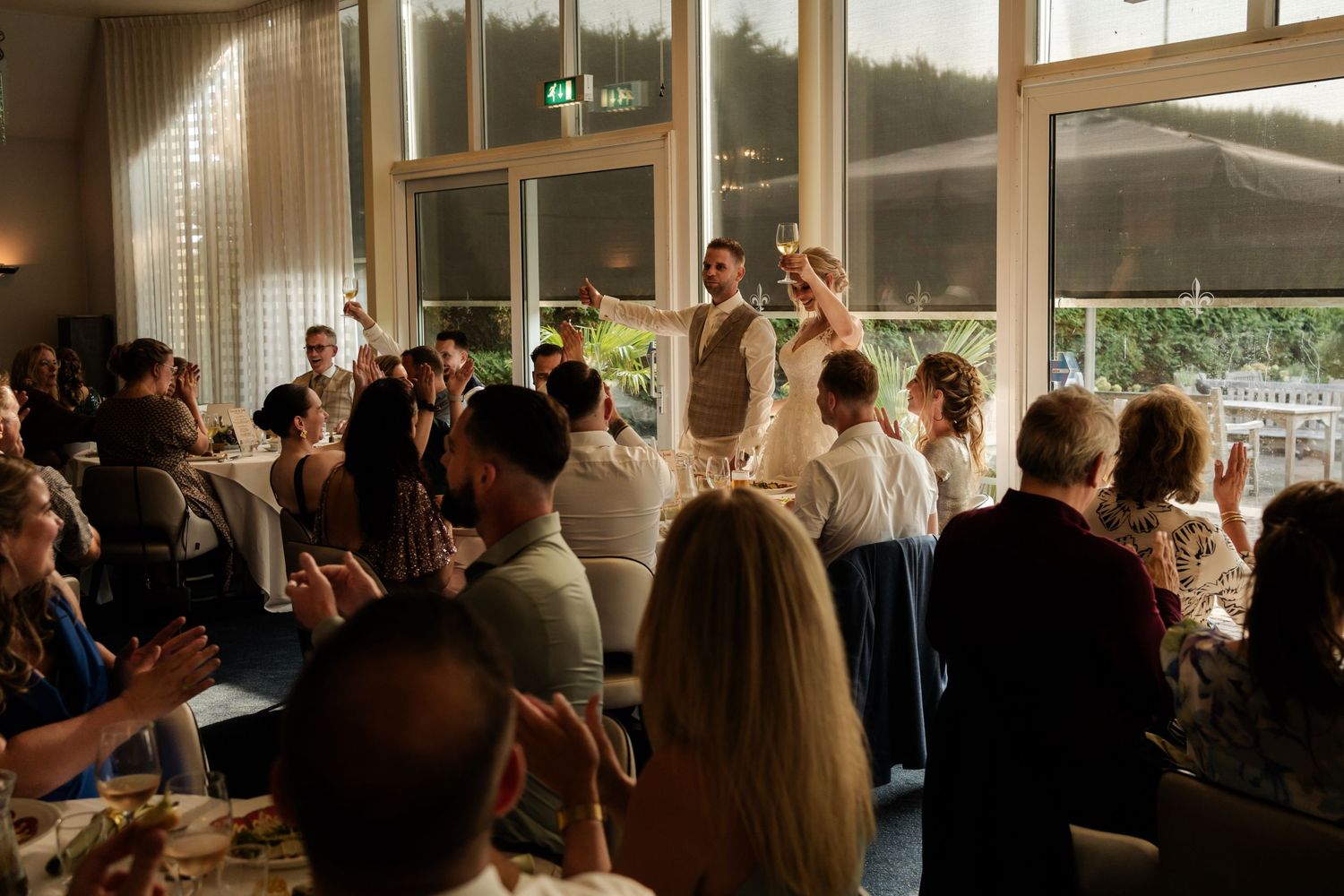 Wedding guests celebrating and dancing during a reception in a venue with large windows at sunset.