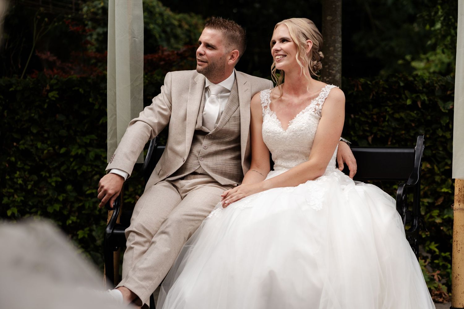 A newlywed couple sits together in formal attire on their wedding day against a garden backdrop.