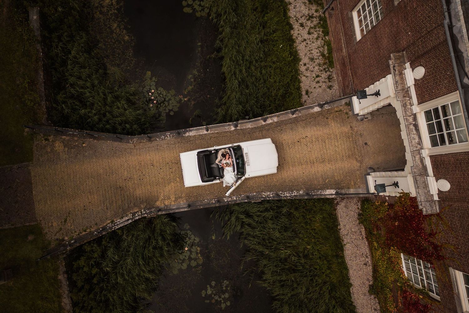 Aerial view of a residential property with a white vehicle parked in the driveway surrounded by trees and landscaping.