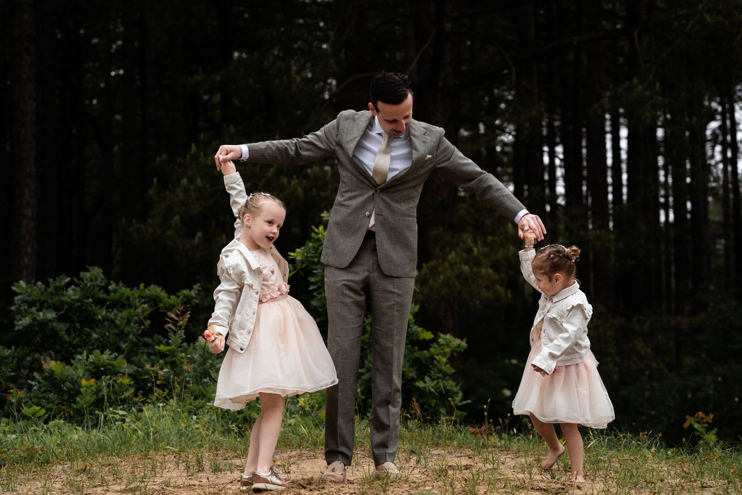 A heartwarming moment as a person in grey suit plays with two young children in pink dresses outdoors.