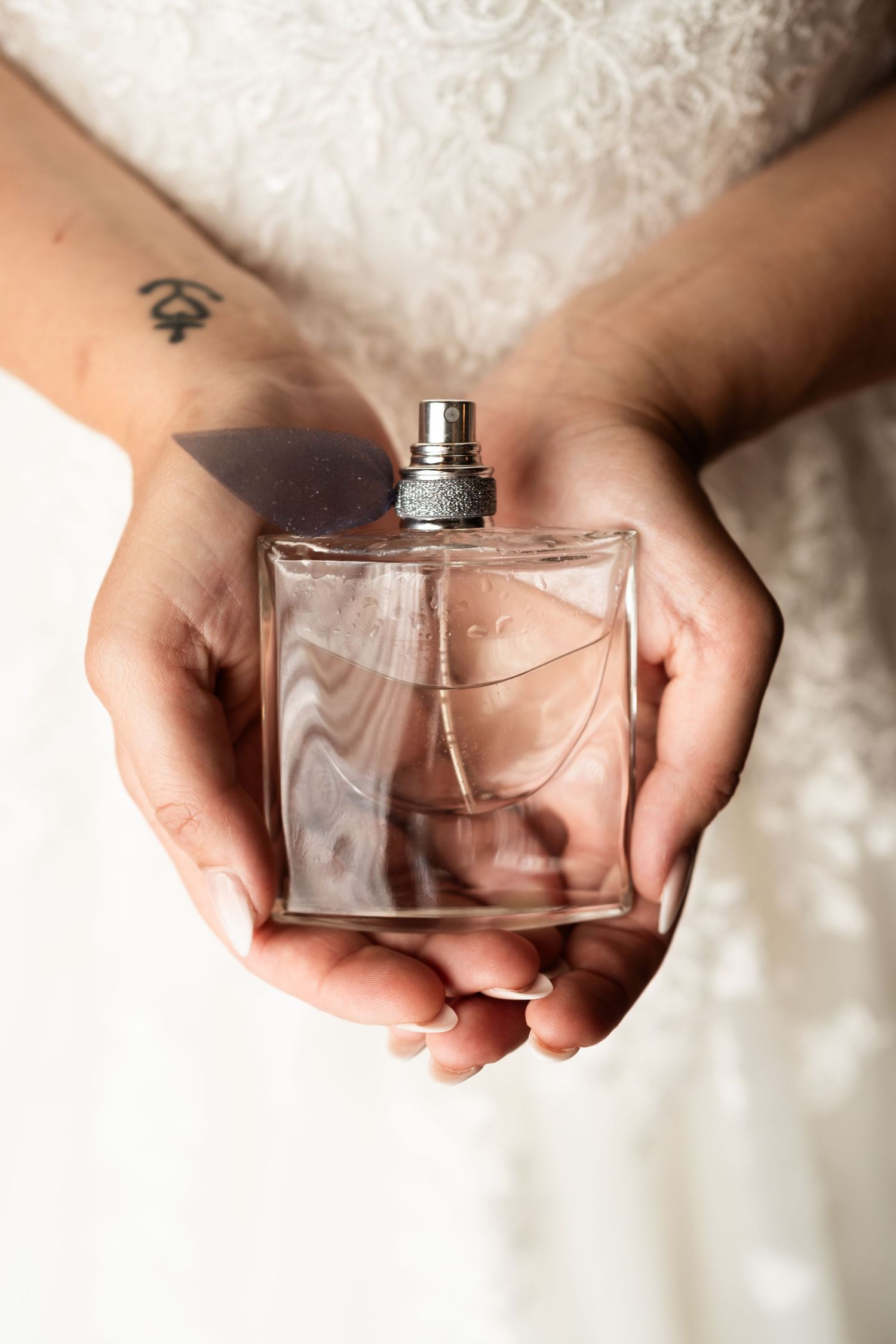 Hands holding a square glass perfume bottle against a white lace dress.