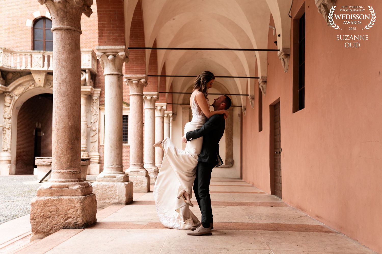 A couple embraces in a romantic dance pose under a beautiful pink arched colonnade walkway.