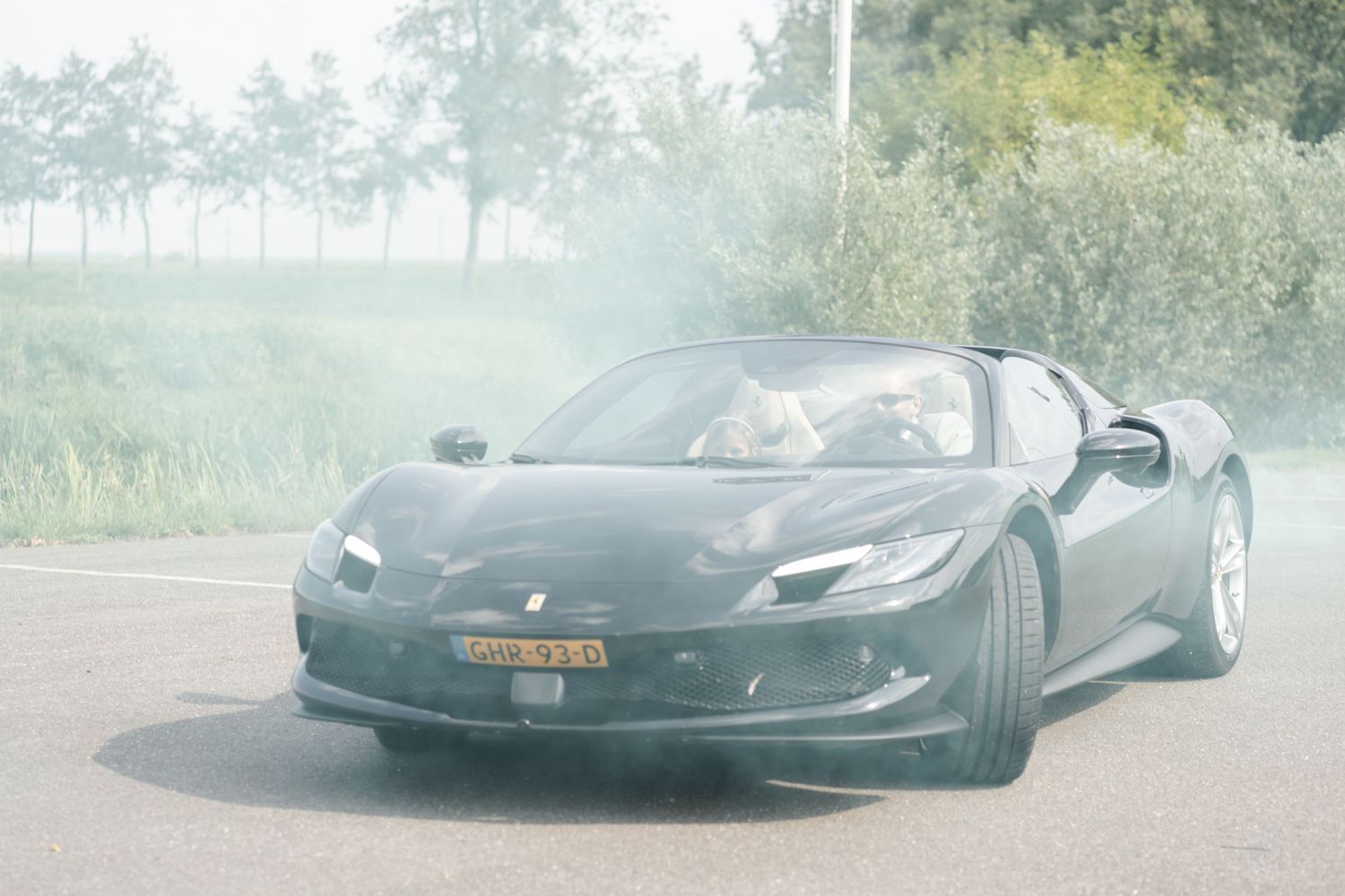 A sleek black Ferrari sports car drives on a misty rural road with trees in the background.