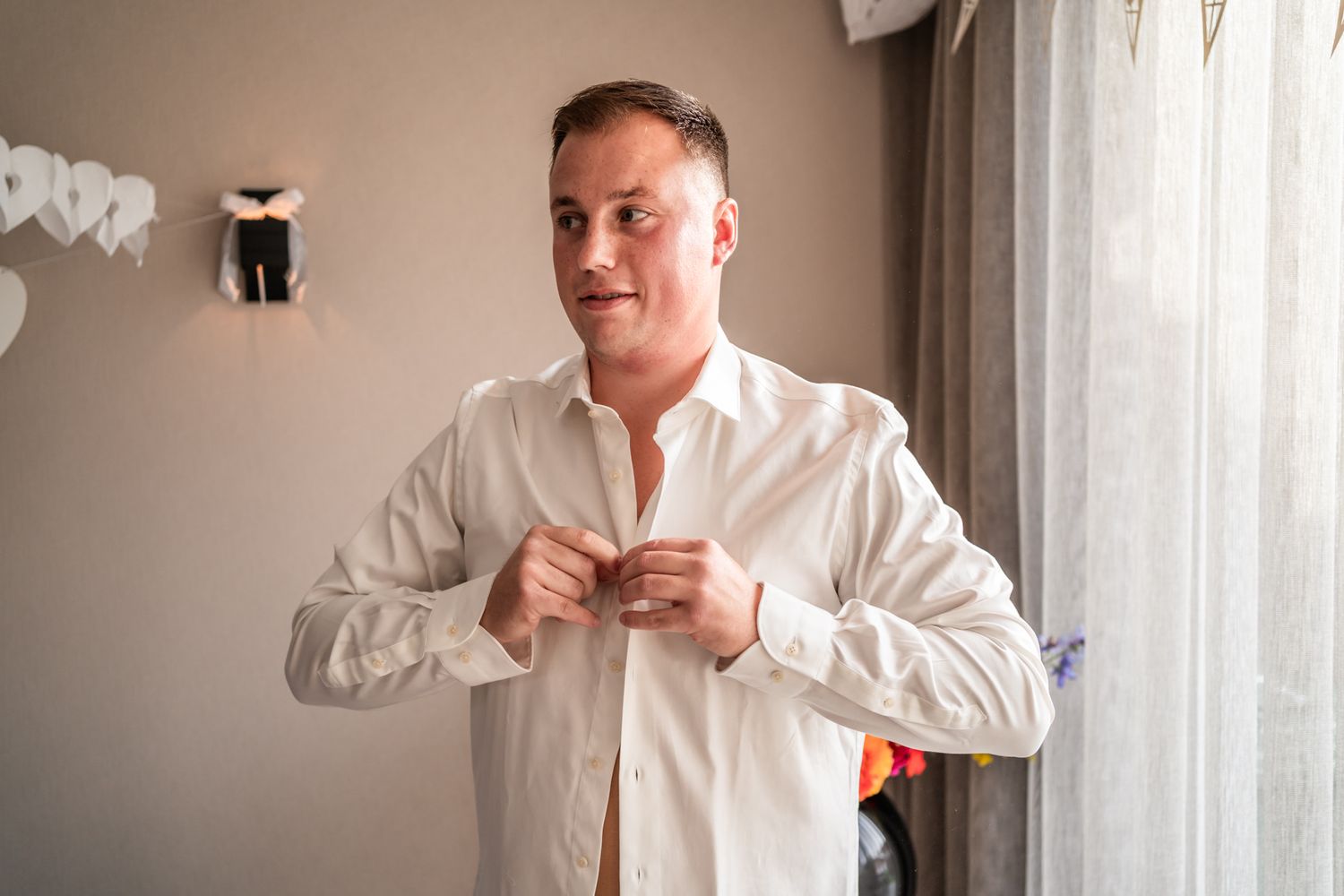 A person in a white dress shirt buttons up while getting ready by a window.