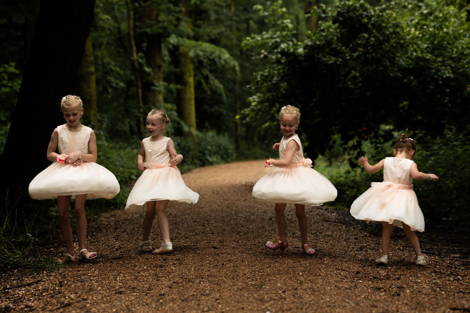 Four young children in white tulle dresses twirl and dance on a forest path.