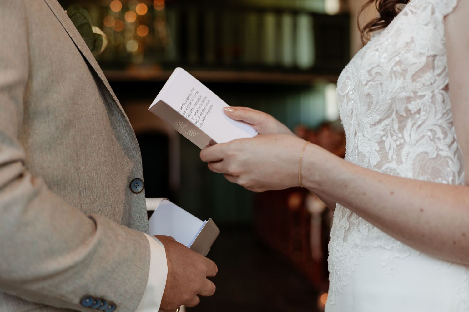 Bride in lace gown reading wedding vows from a card during an intimate ceremony.