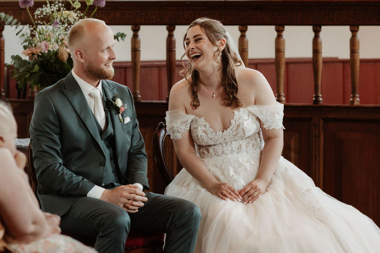 A bride in a floral gown laughs joyfully with her groom in a green suit during their wedding ceremony.