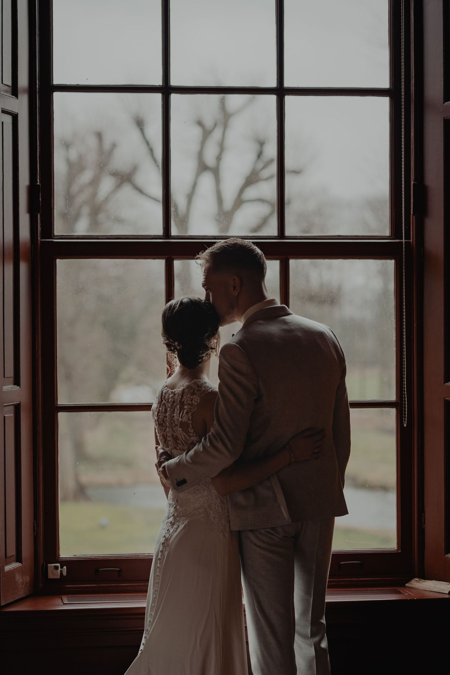 A couple stands together by large windows in a rustic interior space with natural lighting.