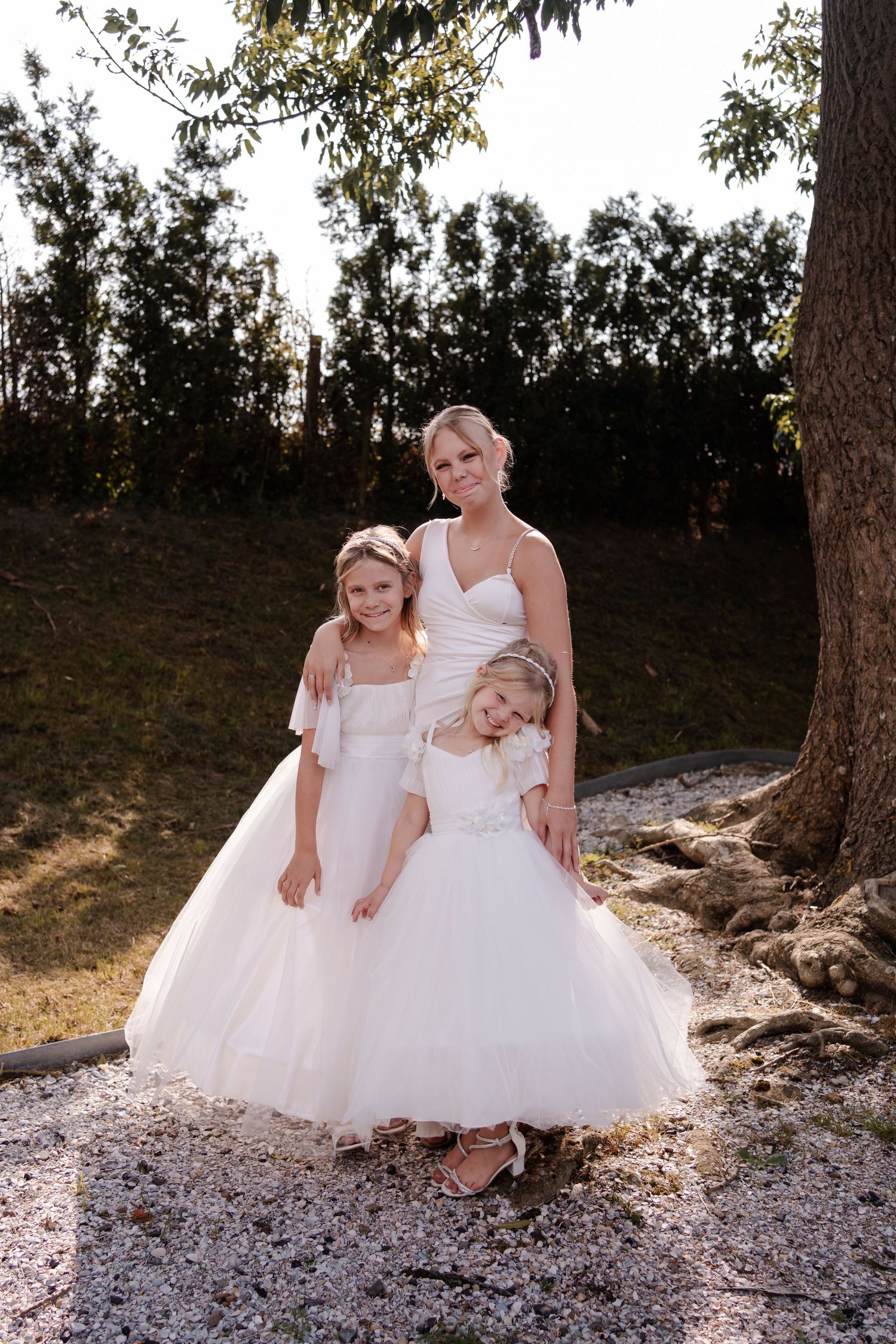 Bride in white wedding dress poses outdoors with two flower girls in matching tulle dresses at sunset.