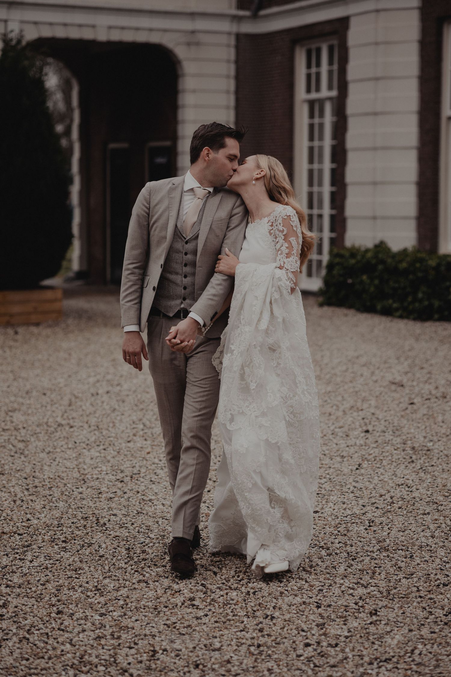 A couple in wedding attire walking together on a gravel path outside an elegant stone building.