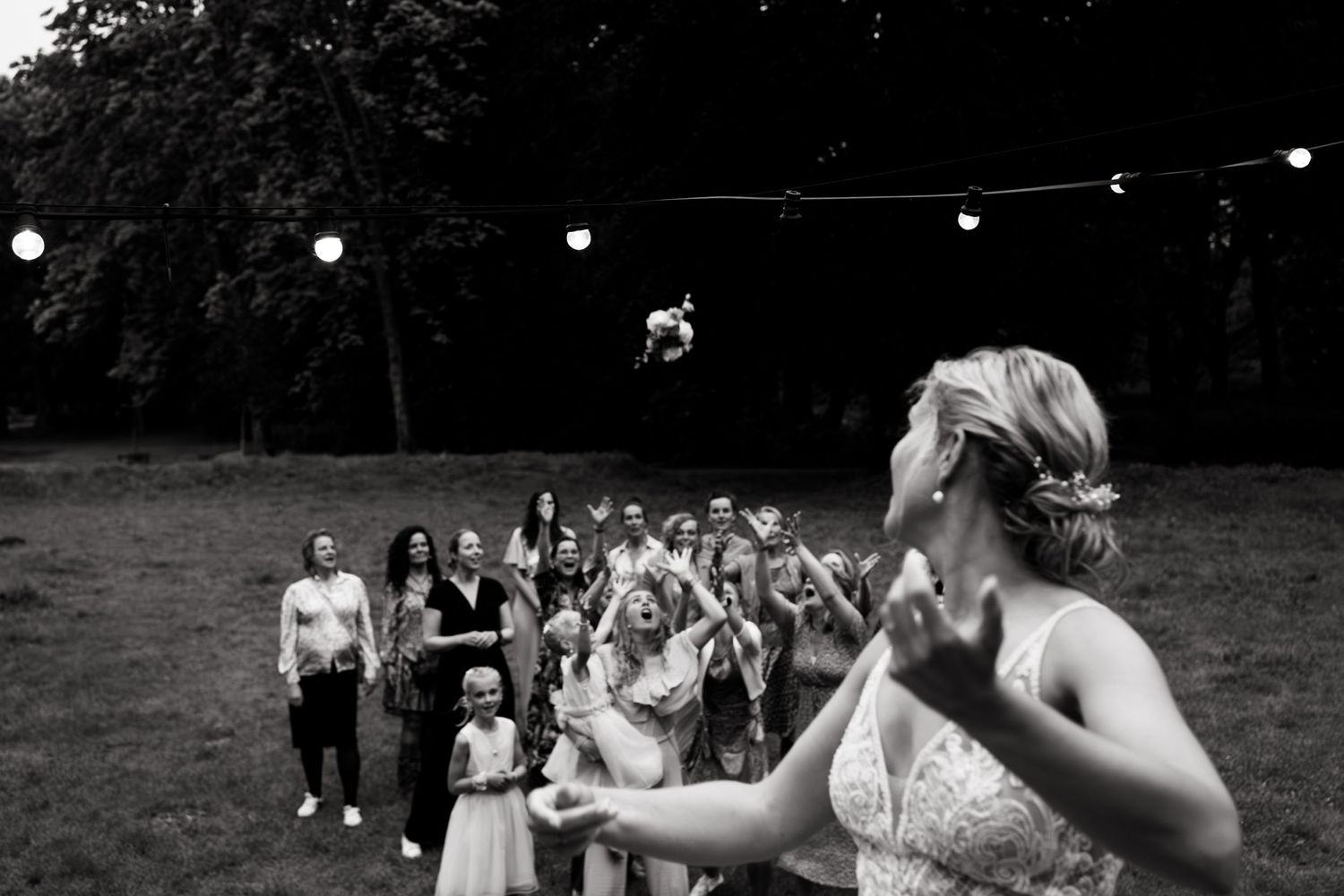 Wedding reception scene with guests gathering for bouquet toss at outdoor evening celebration.